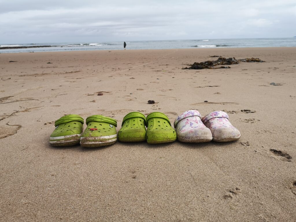 drei paar Crocks Schuhe stehen am Strand und warten auf ihre TrägerInnen