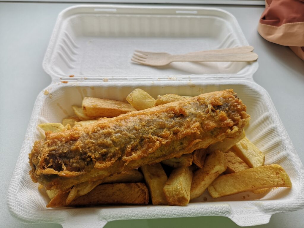 Vor uns liegt eine Portion Haggis udn Chips.Der Haggis ist in einer kleinen Wurstform und außen mit Bierteig umhüllt und frittiert worden. Er liegt oben auf einer Portion schottischer Pommes, die in UK Chips genannt werden.Beides liegt auf einem Fensterbrett in einer to go Pappverpackung in der Chippie Bude