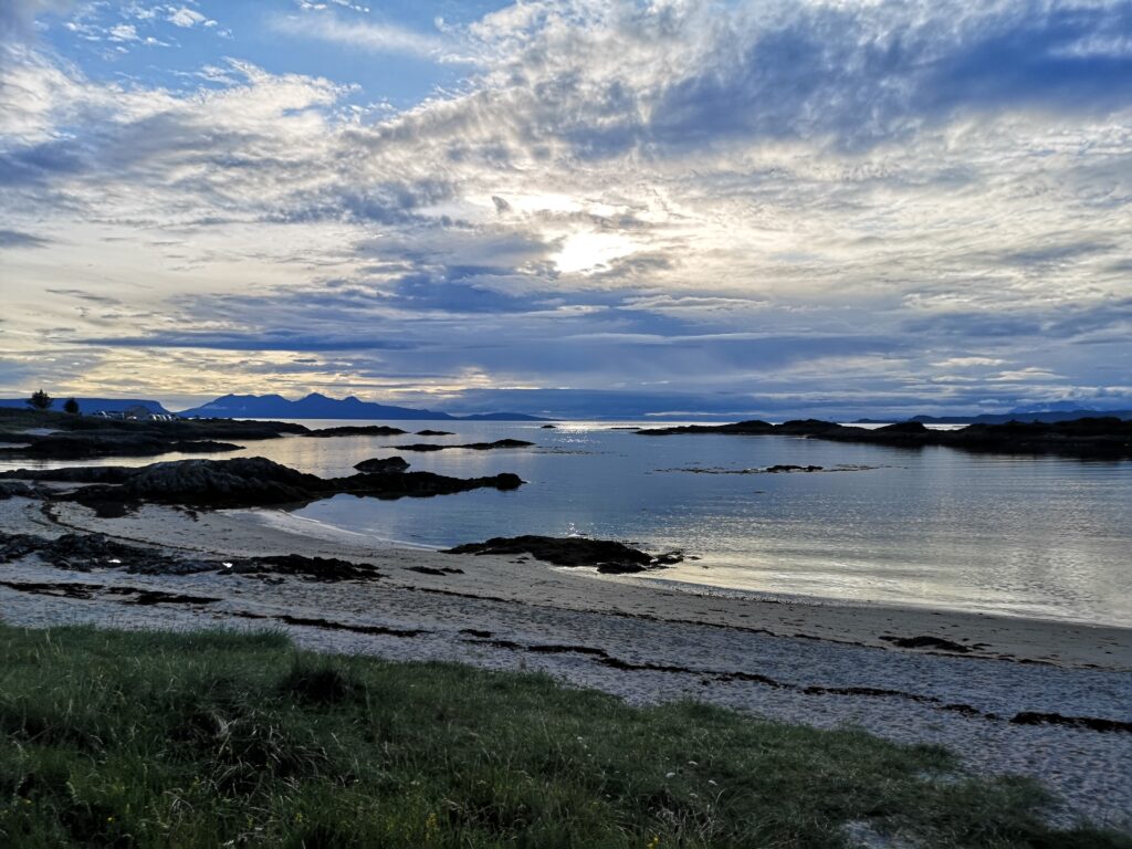 Wir stehen am Ufer am Traigh Golf Course und schauen Richtung Westen.Unter uns ist weißer Sandstand. Die Sonne ist durch Schleierwolken verdekt und beleuchtet die Umgebung indirekt und man sieht leichte Wolken im Meer wiederspiegeln