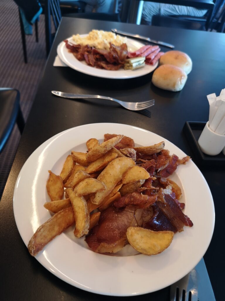 Wir sehen auf eine n Teller der auf einem sehr dunklen Tisch steht. Auf diesem Teller sind gebackene Kartoffelspalten mit gebratenem Speck zu sehen. gegenüber steht ein Teller auf dem gebratene Pilze, gebackene Bohnen in Tomatensoße und Rührei schemenhaft zu sehen sind