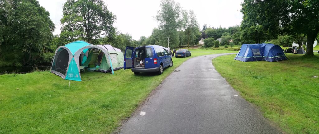 Wir sehe nauf eine Wiese, auf dem wir unser Zelt und Pavillon aufgebaut haben. beides sind in grün gehalten. Daben steht unser Urlaubsauto, ein blauer VW Caddy.Unser Zeltplatz und wo wir das Zelt aufgebaut haben, ist direkt an einem Fluss, und auf der anderen Seite von Bergen begrenzt. Unser Stellplatz ist direkt am Fluss