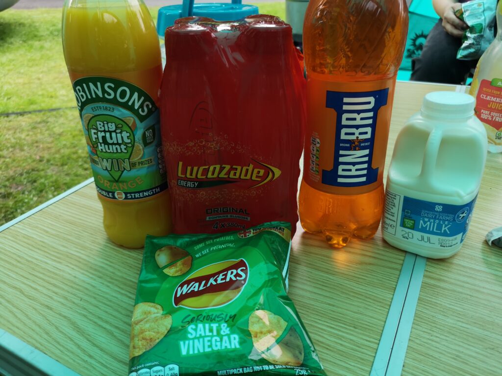 Auf einem Campingtisch in Holzoptik habe ich einen Teil unseres ersten Einkaufs aufgebaut.Darunter sind "Robinsons Squash", ein orangensirup den ma nmit Wasser verdünnt. Dann ein 4er Pack Lucozade und eine Flasche IrnBru, das sidn zwei Energie Driks in Schottland, davor liegt eine Tüte Warkers Chrisp, als Kartoffelchips in Geschmacksrichtung Salz und Essig