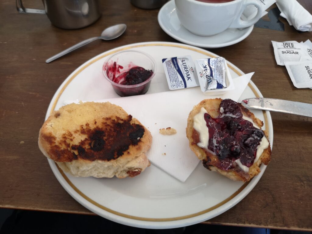Auf einem Tisch steht ein Teller, auf dem aufgeschnittene Fruitescones liegen. Diese sind innen noch geröstet worden. Auf einer Hälfte wurde schon Butter und Marmelade gestrichen und wartet darauf gegessen zu werden