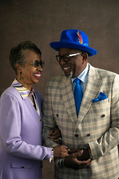 Alex and Karen Brown stand smiling ad hooking arms in a lavender blazer and checkered blazer with blue accoutrements