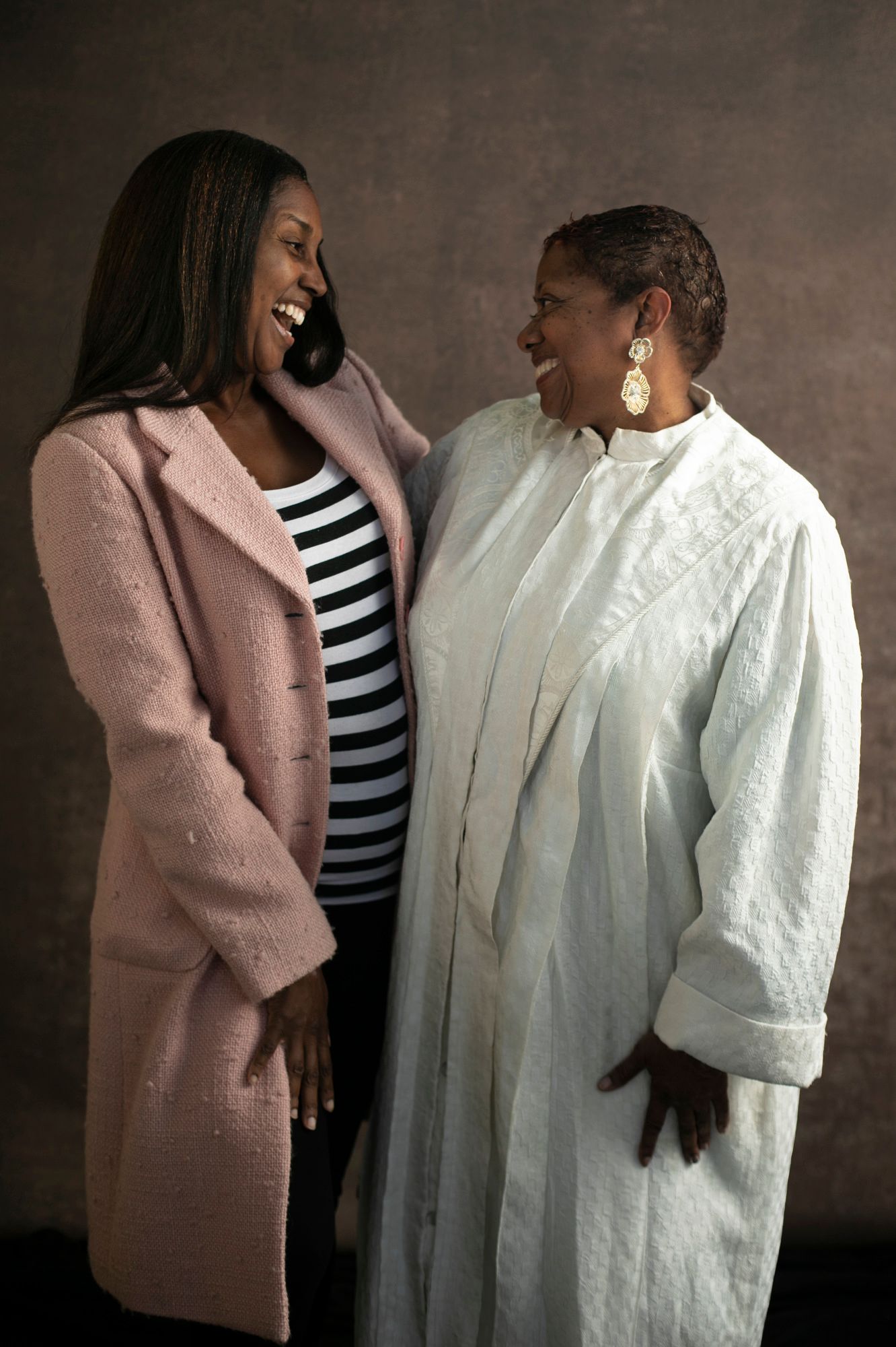 Danielle Wortham and Reverend Judy Wortham, laugh toward each other as they embrace in a pink trenchcoat and white robe, respectively