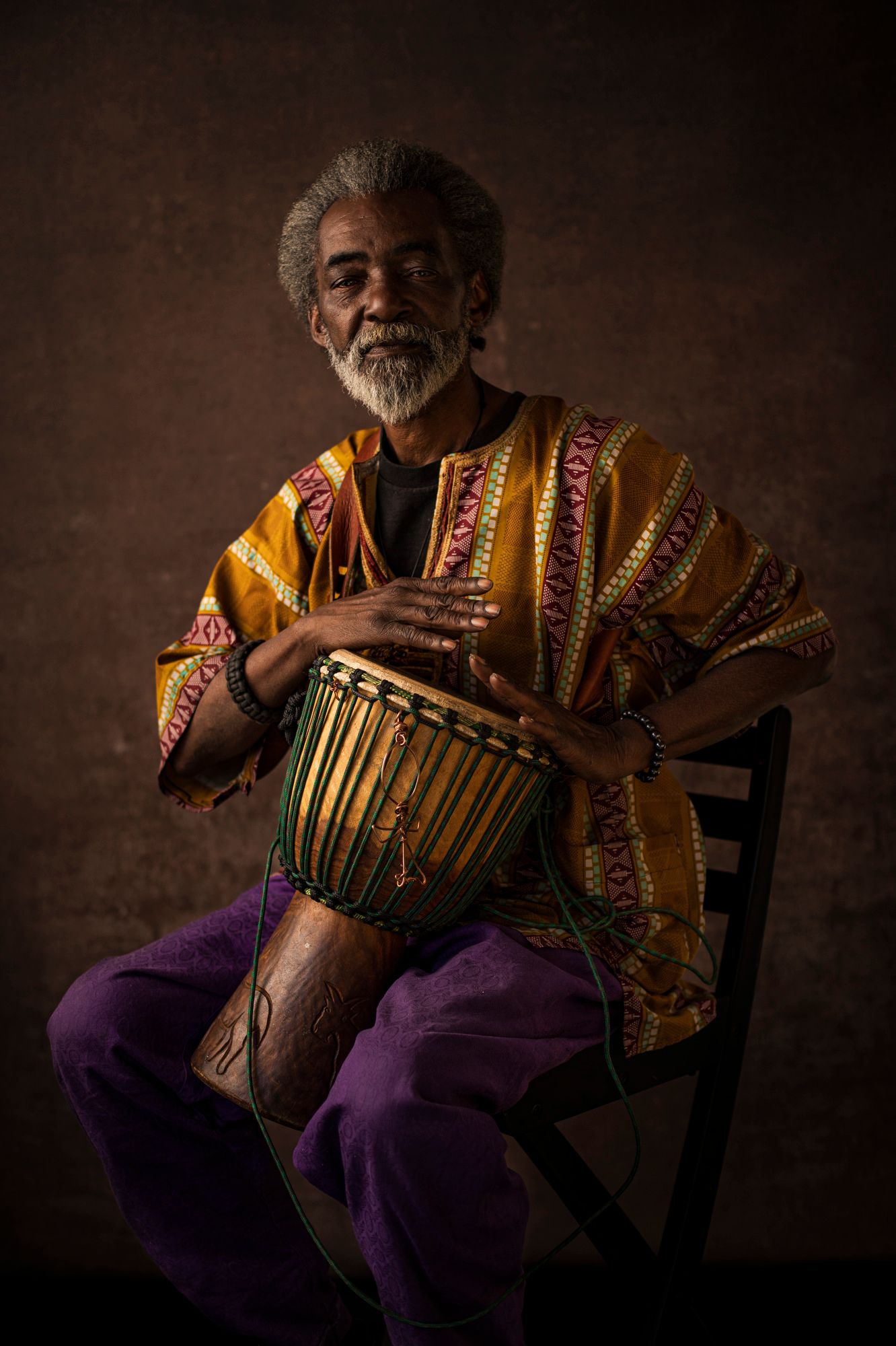 Roi-el sits in a bright shirt and pants with his hands mid-stroke on a drum
